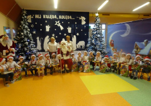 Grupa dzieci z prezentami, pani dyrektor Maria Królikowska, pani Agnieszka Piekarska oraz Mikołaj na tle świątecznej dekoracji.